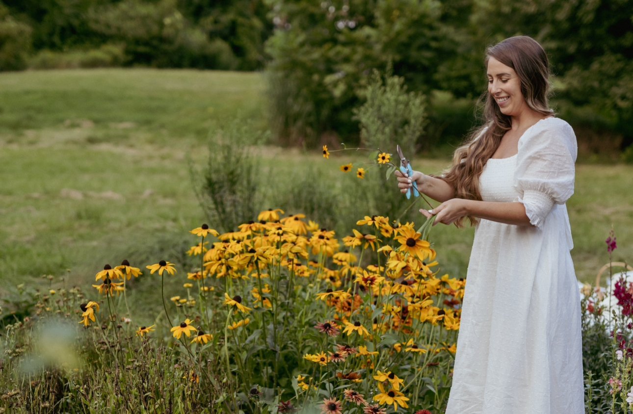 A woman in a white dress is smiling while cutting yellow flowers in a garden.
